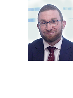 Peter Kohlhagen wears a dark suit and maroon polka dot tie, smiling against a bright, blurred background.