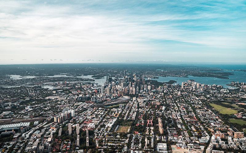 Aerial view of a dense city skyline with high-rise buildings, surrounded by water and sprawling suburbs under a partly cloudy sky.